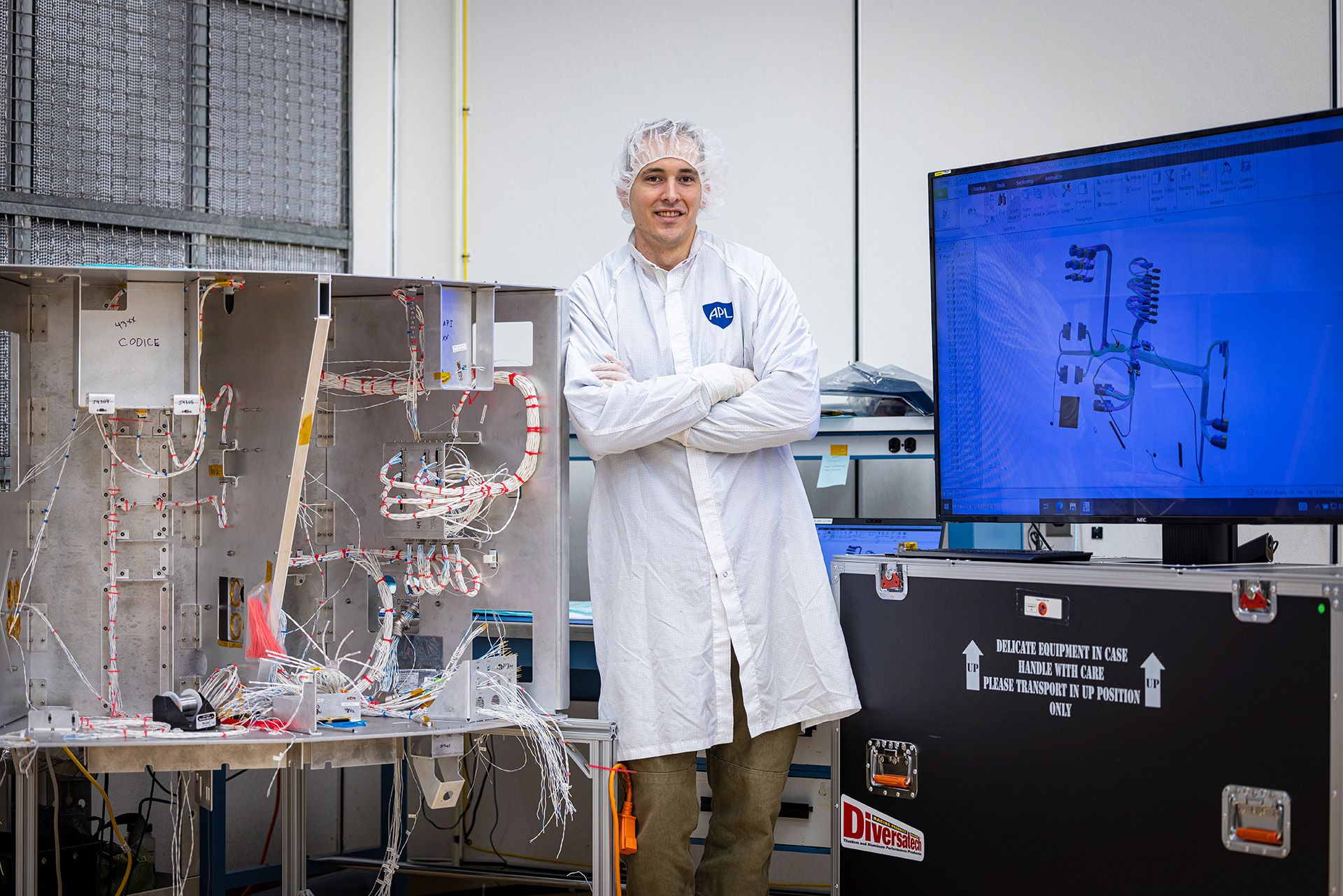 APL engineer Kyle Norman stands between a computer-aided design (CAD) drawing of the Interstellar Mapping and Acceleration Probe (IMAP) wiring harness and the harness itself on a mock-up of the spacecraft.