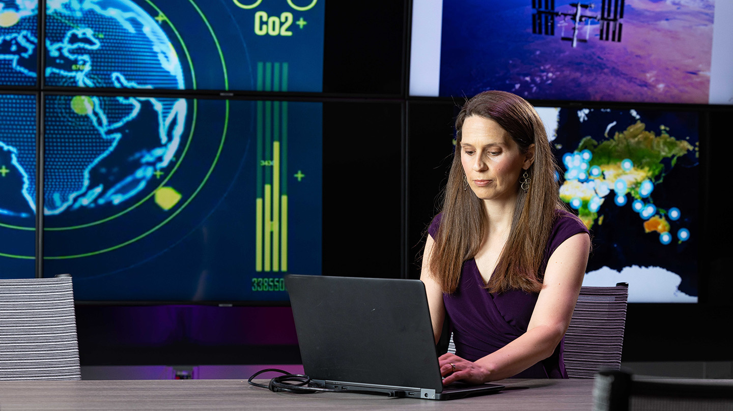 Elizabeth Reilly types on a laptop. In the background, screens display greenhouse gas emissions data.