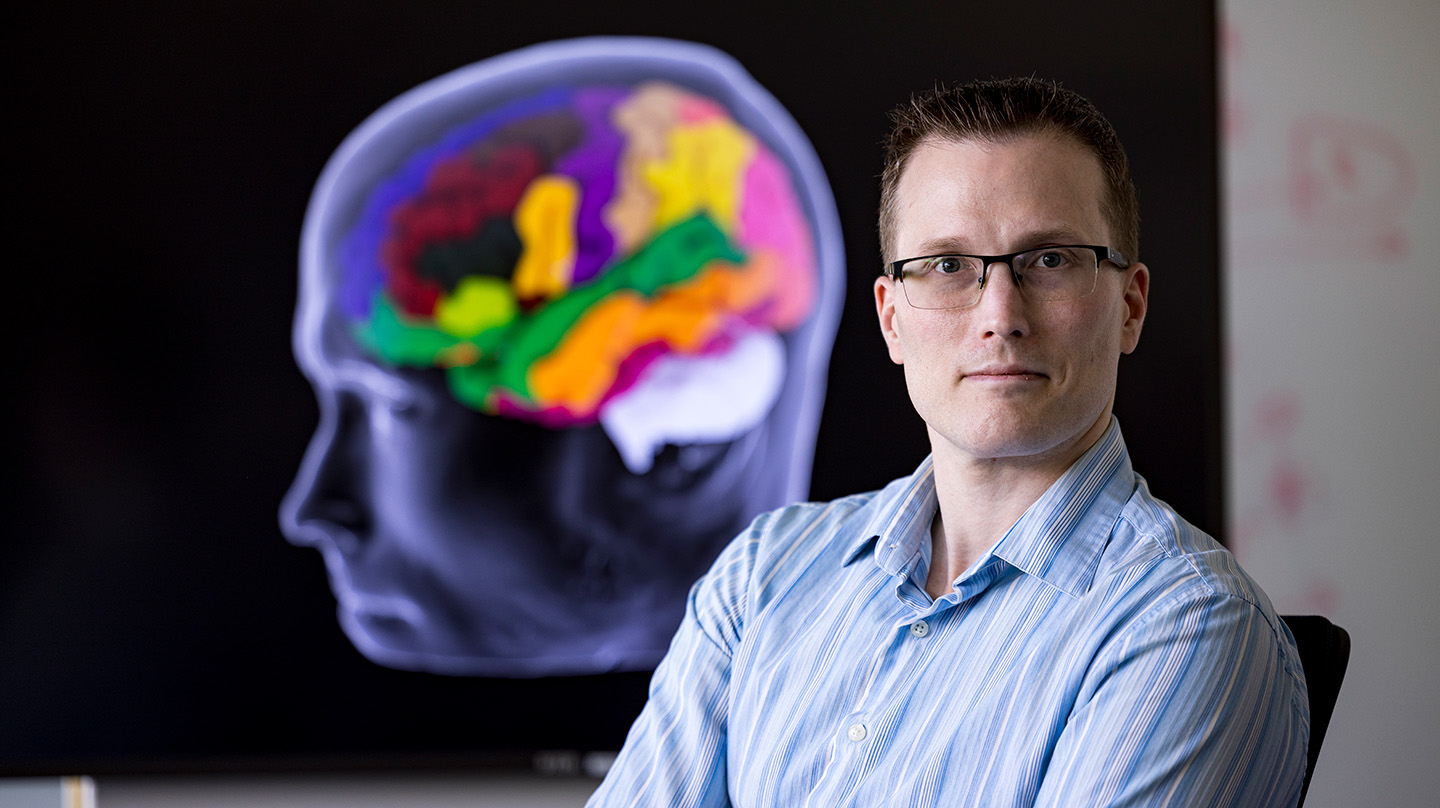 Will Coon sits in front of a 3D rendering of the human brain