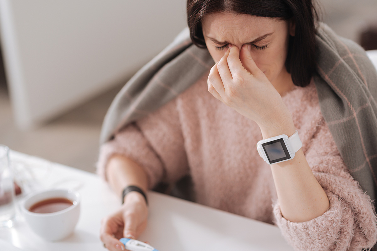 A person with a cold holds a thermometer. A wearable device is on their wrist. (Credit: Bigstock)
