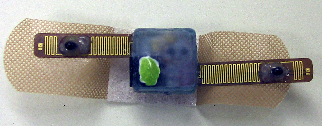 The underside of a prototype Ambulatory Raynaud's Monitor, approximately 9/16 of an inch in length, is shown on top of a Band-Aid, which is often used to secure the monitor to a patient's finger